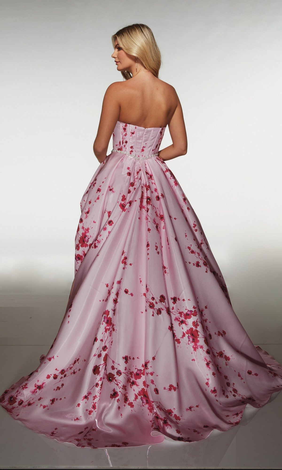 A woman wearing the Alyce 61989 long prom dress stands facing away, highlighting the strapless satin floral gown’s corset bodice and flowing skirt.